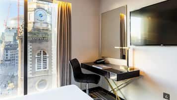 A hotel room with a desk, chair, and a large TV. There's a window with a view of a building and a clock.