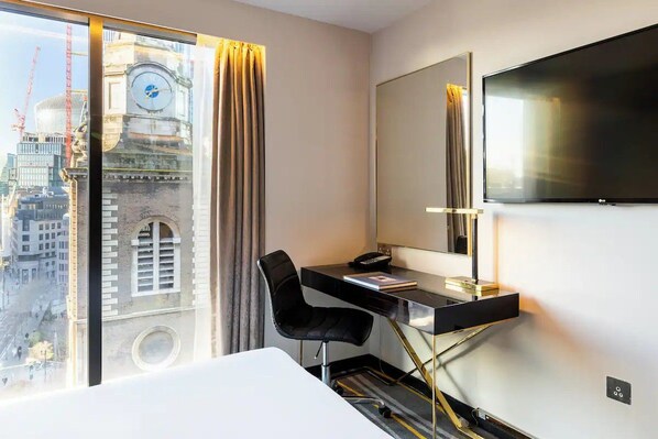Hotelkamer met een bureau, stoel en een grote televisie. Een raam biedt uitzicht op een gebouw en er hangt een klok.