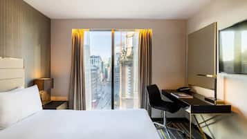 A hotel room with a bed, a desk, a chair, a TV, and a large window with curtains.