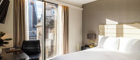 A hotel room with a large bed, a desk, a chair, a lamp, and a view of the city through a large window.