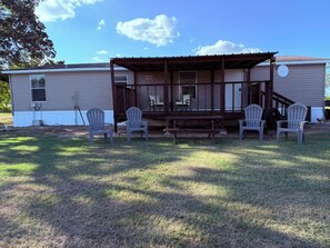 A covered outdoor seating area with four chairs and a table, a mobile home in the background, and a clear sky.