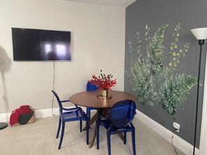 Een eethoek met een ronde tafel, blauwe stoelen, een flatscreen televisie aan de muur en een muurschildering van begroeiing.