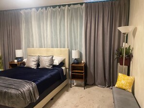 Een slaapkamer met een bed, twee nachtkastjes, een lamp, een bankje en gordijnen.