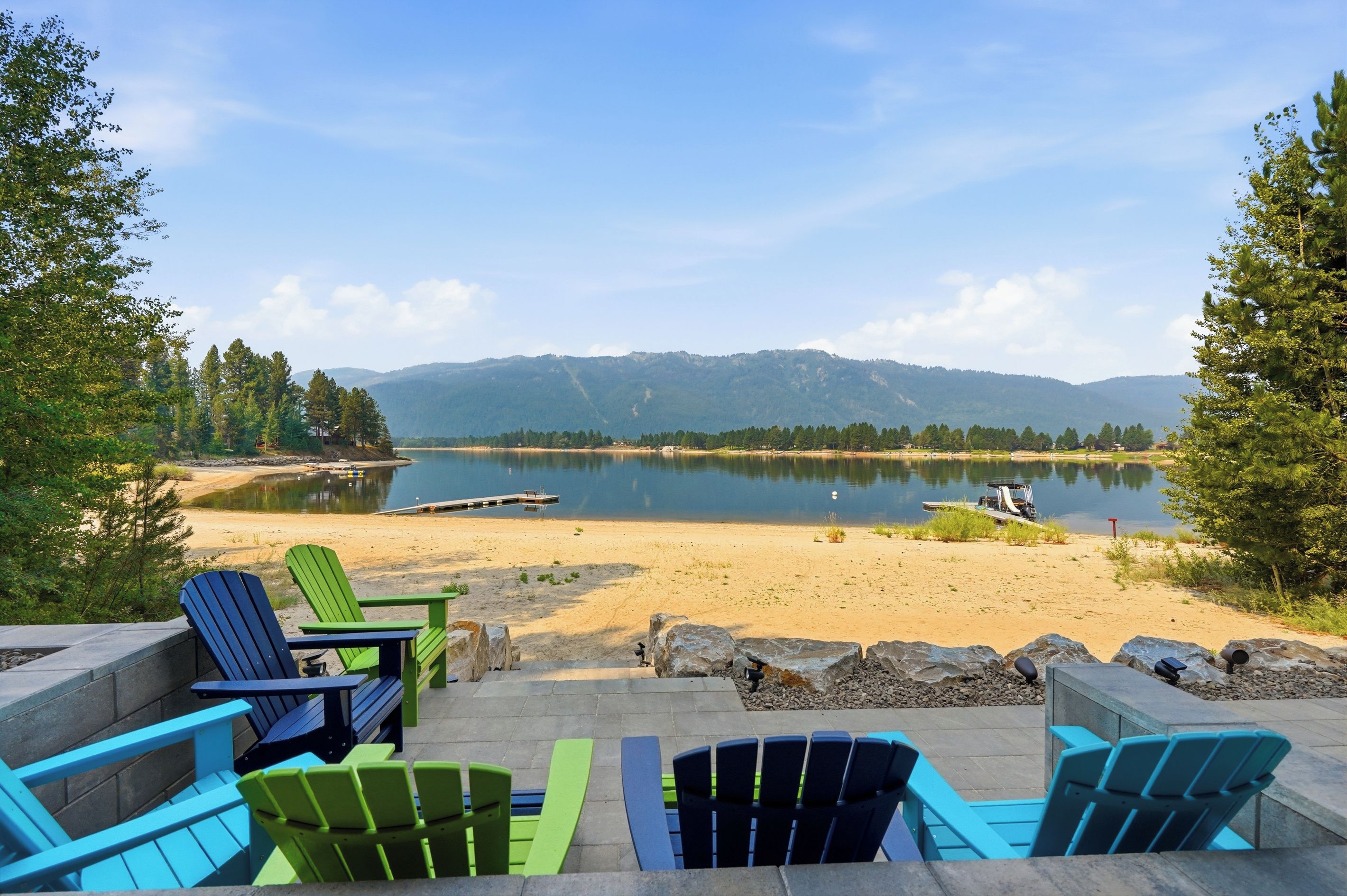 Un espacio de descanso junto al lago con sillas Adirondack y un pozo de fuego, con vistas a un lago tranquilo y montañas lejanas.