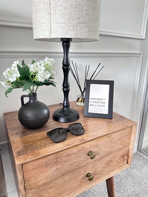 A wooden nightstand with a lamp, a vase with white flowers, a framed inspirational quote, and a pair of sunglasses. - Stylish Central Derby 2 Bed, 2 Bath | Free Parking, Gym & Near Station (Derby)