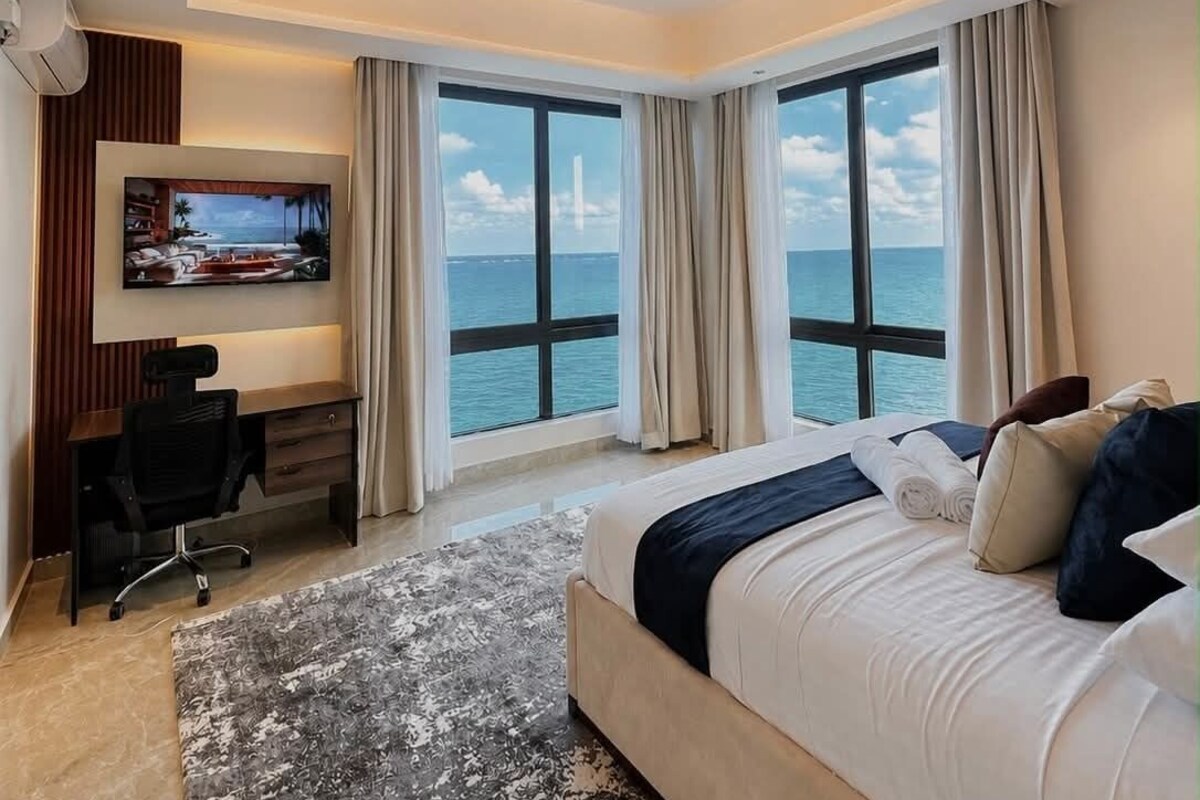 Habitación de hotel con cama, escritorio, silla, televisor y ventanas grandes con vista al mar.