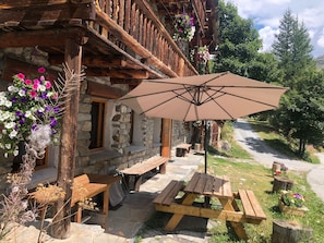 Ein rustikales Steinhaus mit hölzernen Balkonen, Außenbestuhlung und einem großen Sonnenschirm, der Schatten spendet.