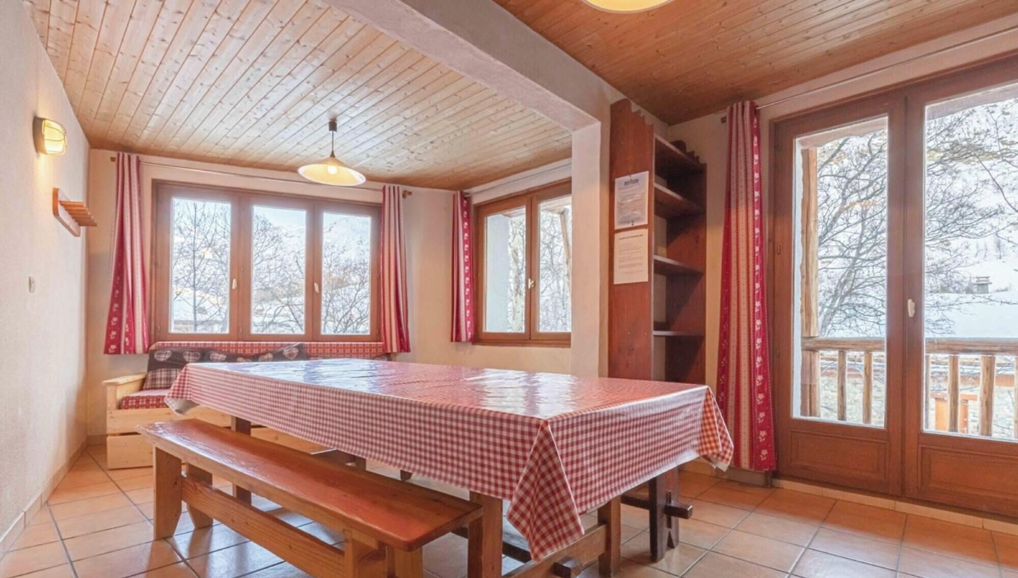 A room with a wooden table and benches, large windows, and a ceiling with wooden beams.