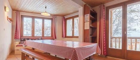 A room with a wooden table and benches, large windows, and a ceiling with wooden beams.
