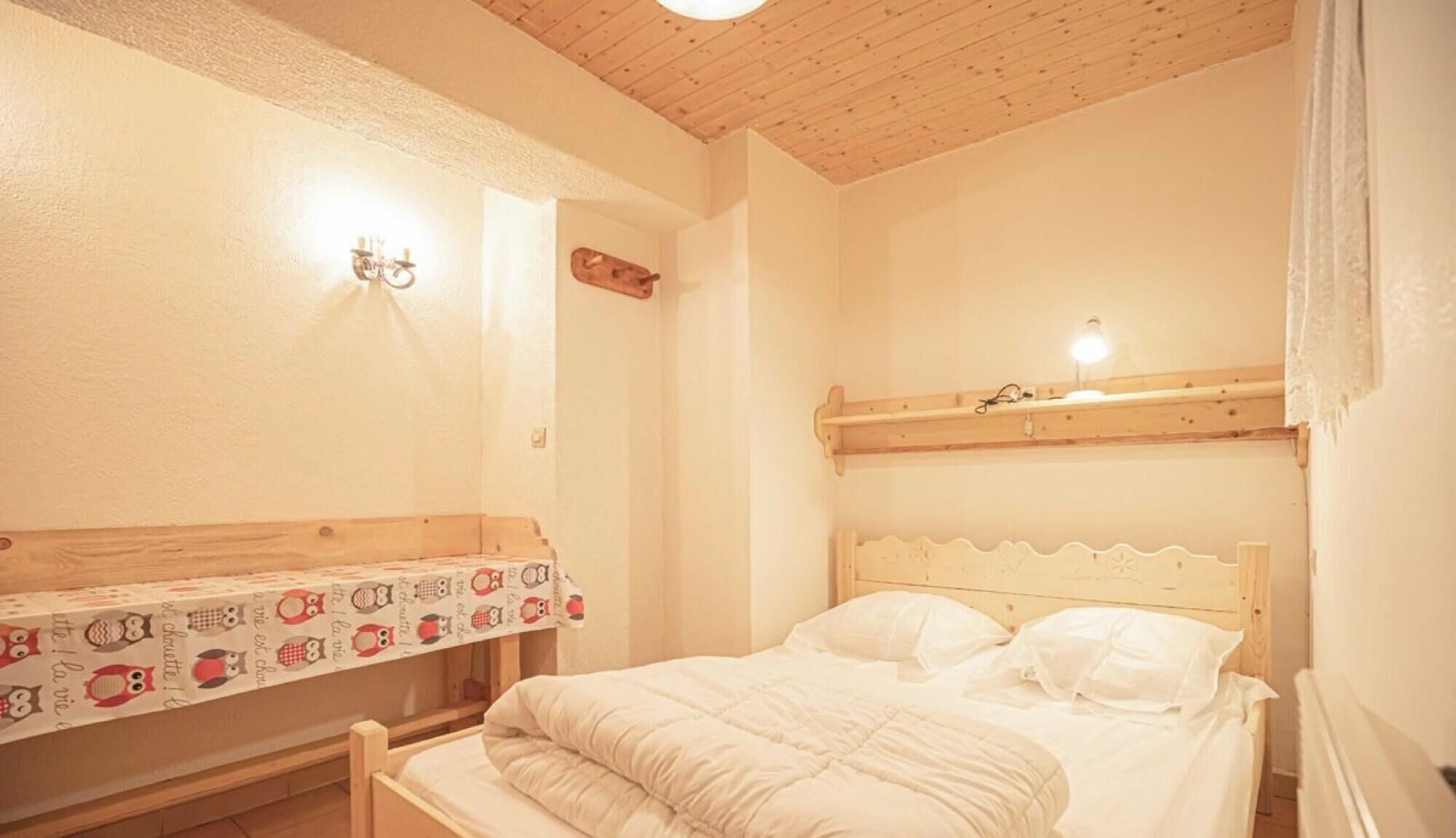 Un lit simple avec une tête de lit blanche et une couvre-lit à motifs, un banc en bois et une étagère murale avec une lampe.