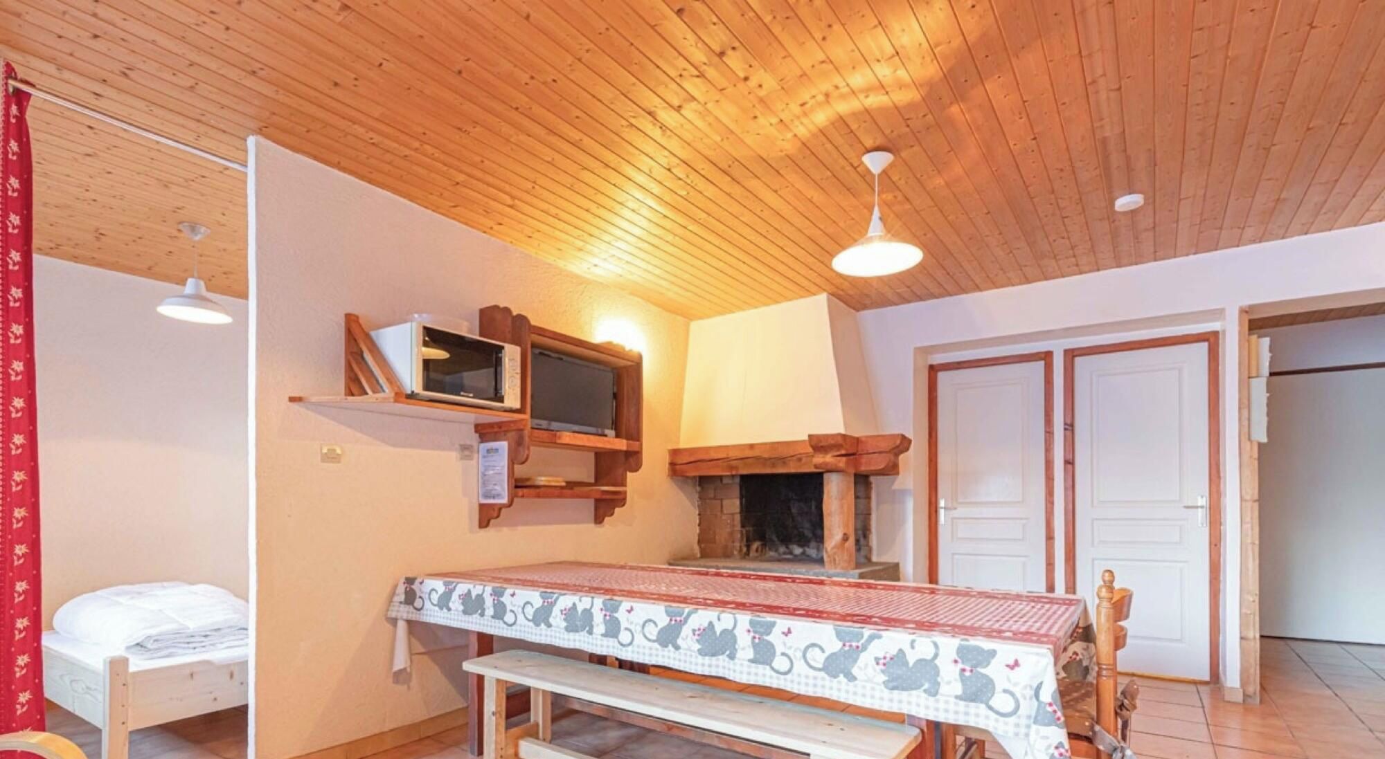 Ein gemütliches Zimmer mit Holzbalkendecke, Kamin, einem Bett, einem Tisch mit einer katzenmusternden Tischdecke und einem Fernseher auf einem Regal.