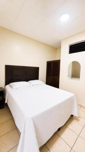 Un dormitorio con cama, mesita de noche, armario y ventana pequeña.
