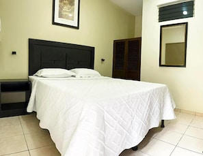Una cama bien hecha con cubrecama blanco en una habitación con suelo de baldosas, cabecera oscura y un cuadro en la pared.