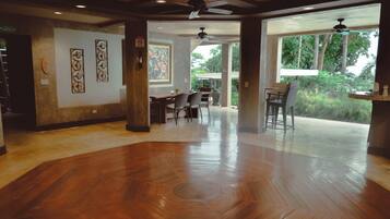 A spacious room with a wooden floor, a dining area with chairs and tables, and large windows offering a view of greenery outside.