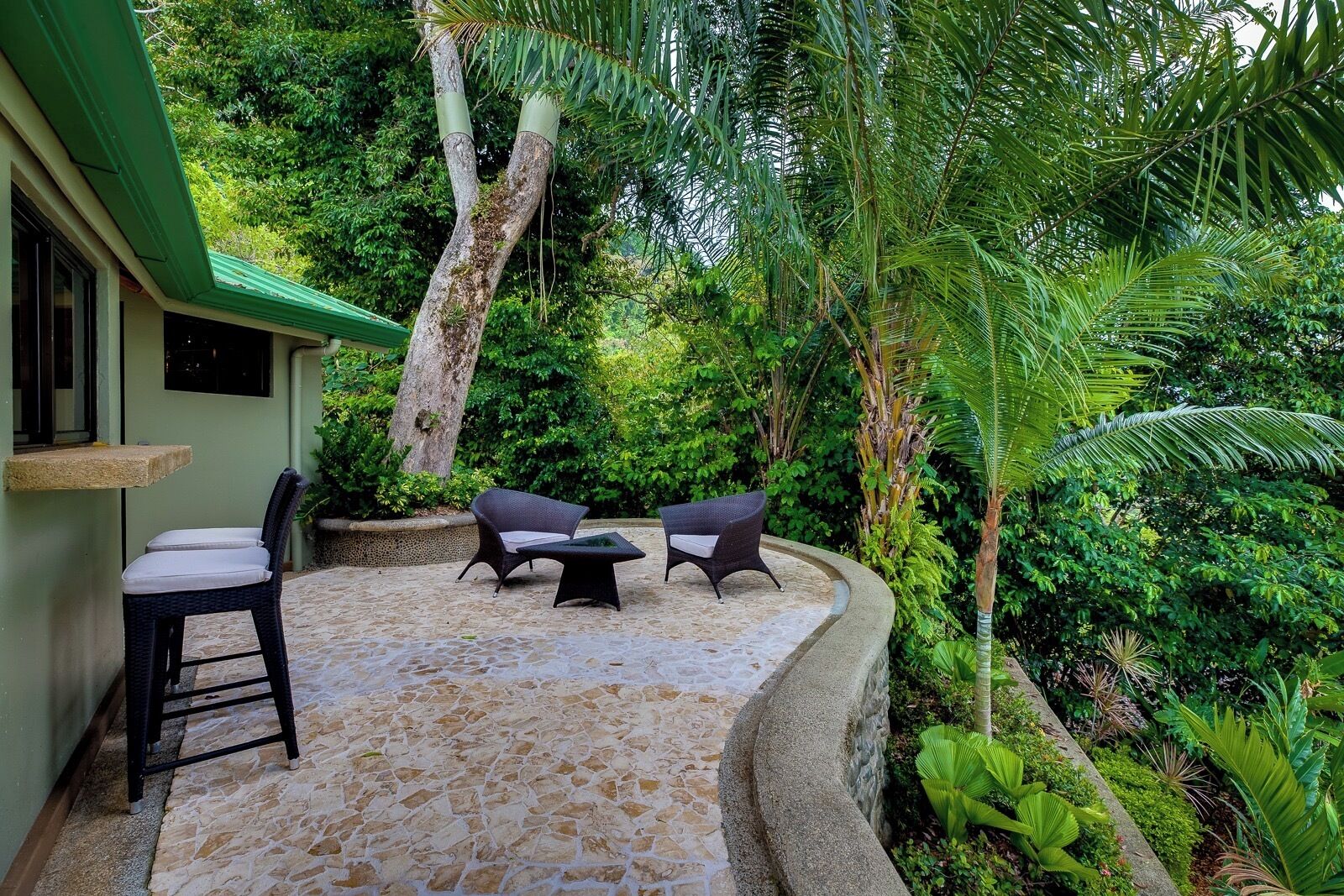 An outdoor seating area with two chairs and a table, surrounded by lush greenery and a stone-tiled floor.