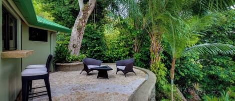 An outdoor seating area with two chairs and a table, surrounded by lush greenery and a stone-tiled floor.