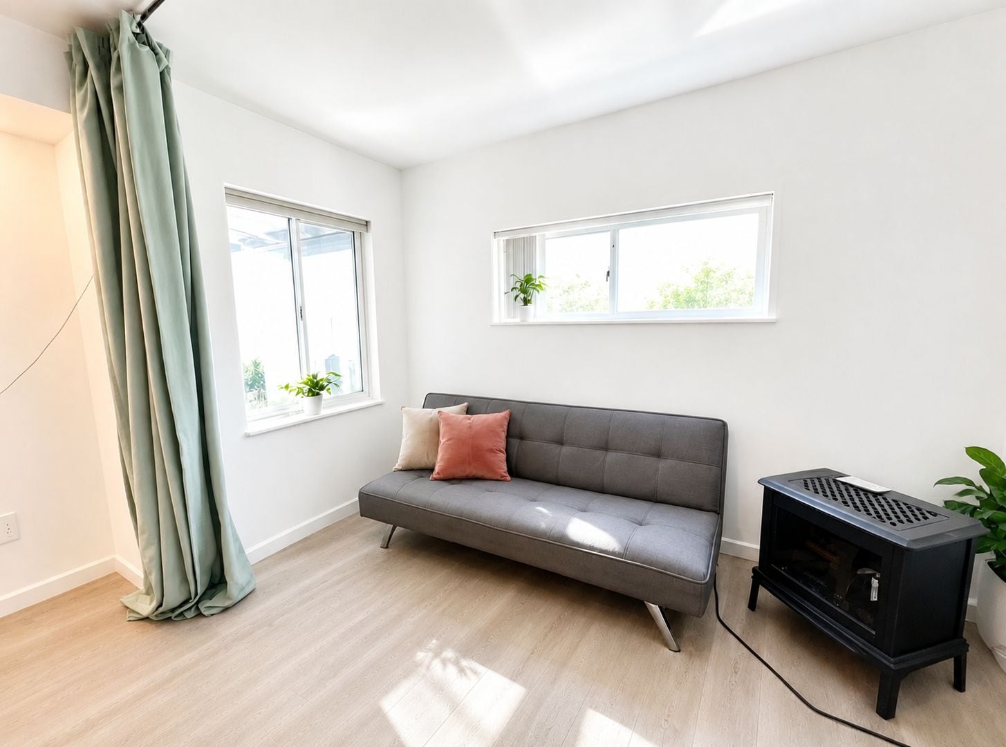 Una sala de estar moderna con un sofá gris, dos ventanas, un calentador negro y una cortina verde.