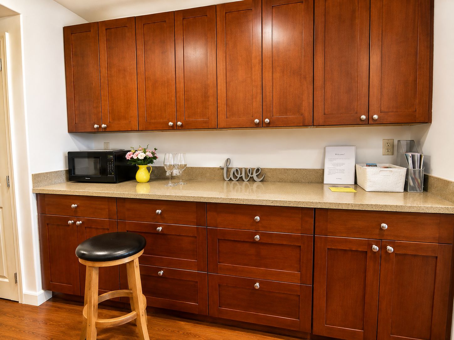A kitchen with wooden cabinets, a microwave, a vase with flowers, and a stool.