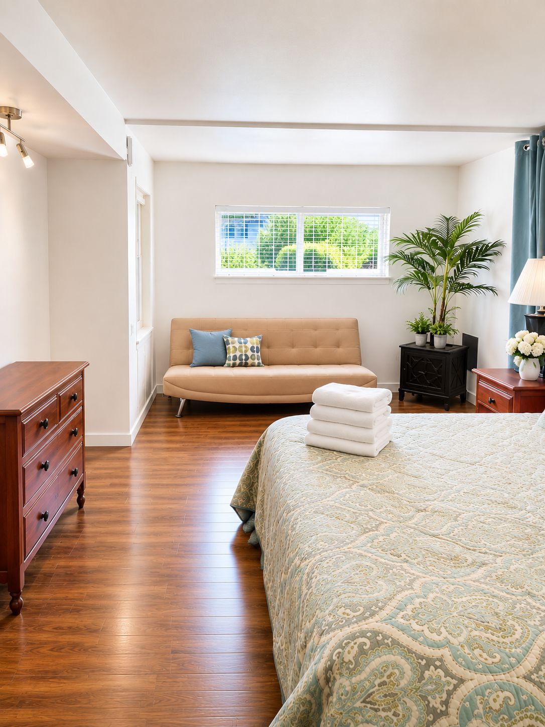 A bedroom with a bed, a sofa, a dresser, a window with a view, and a potted plant.