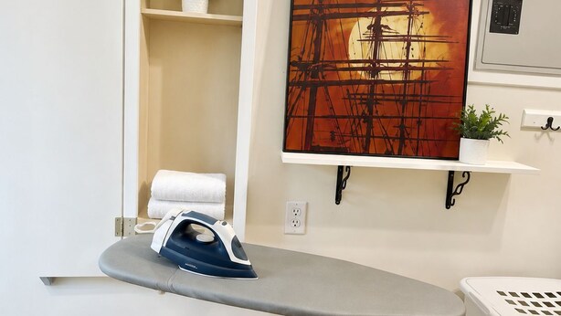 A room with a built-in ironing station, a wall-mounted shelf with a plant, a power outlet, and a laundry basket.
