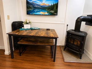 A wooden dining table with a bench, a black wood-burning stove, and a TV mounted on the wall displaying a mountain landscape.