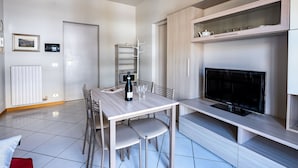 Un comedor moderno con una mesa, sillas, una botella de vino y copas. A la derecha, hay un televisor sobre un mueble.