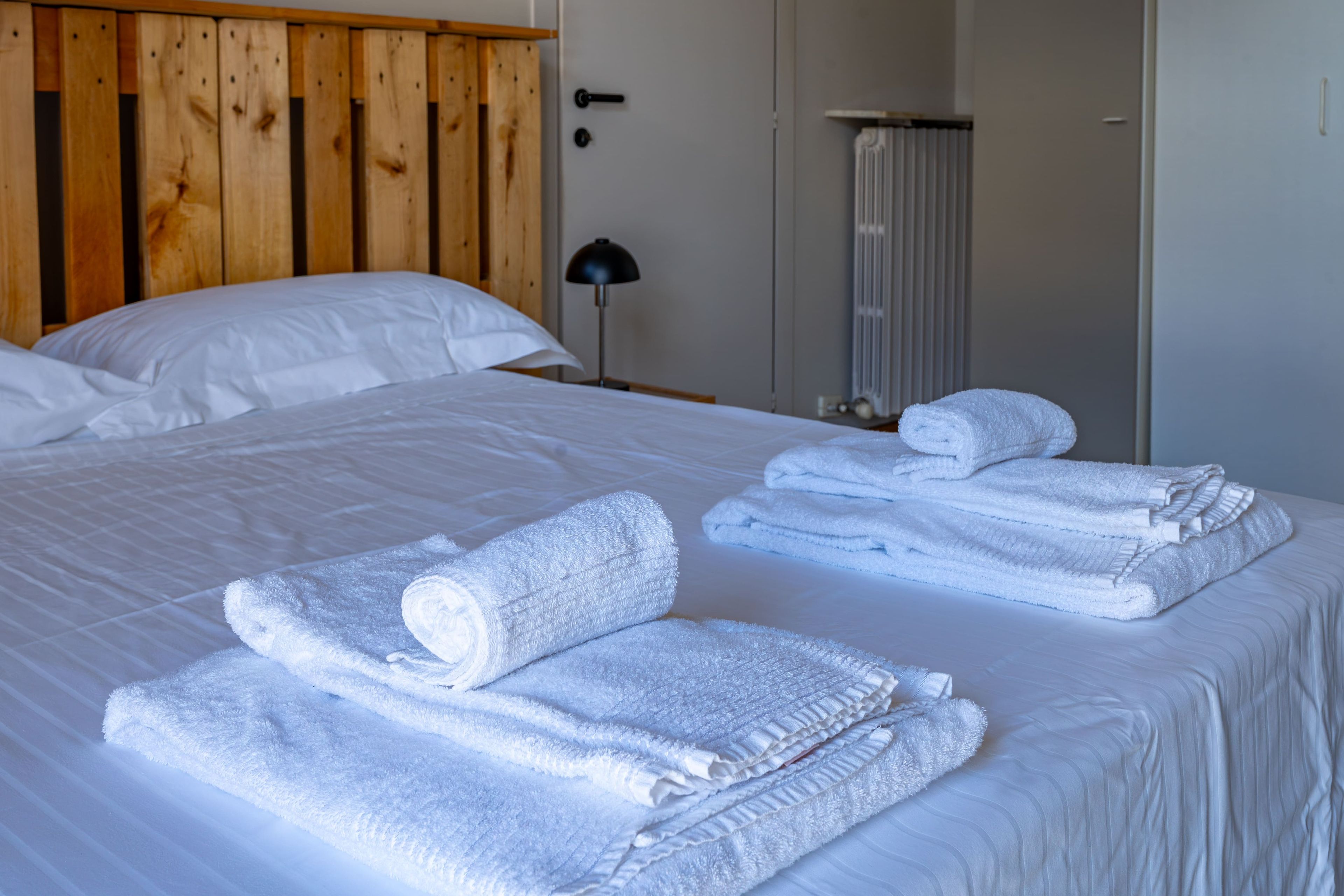 Una cama bien hecha con sábanas blancas y toallas enrolladas, cabecera de madera, lámpara de noche y radiador.