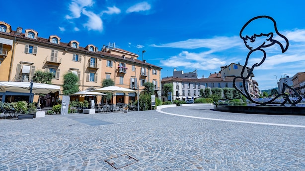 Una plaza empedrada con mesas al aire libre, edificios con balcones y una escultura.