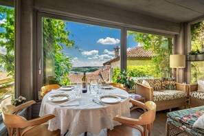 A dining area with a round table set for four, surrounded by wicker chairs, with a view of a tiled roof and greenery outside.