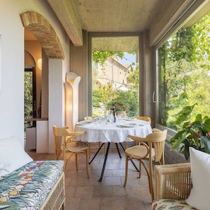 A dining area with a table set for four, surrounded by wicker chairs, a cushioned bench, and a large window offering a view of greenery and buildings.