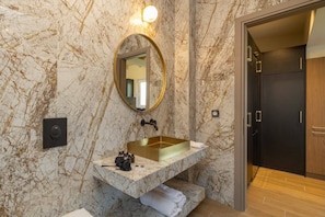 Un baño con pared de mármol, espejo con marco dorado, lavamanos moderno y puerta oscura.