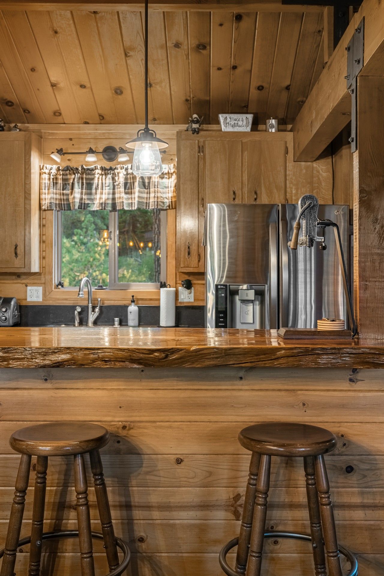 A wooden kitchen with a stainless steel refrigerator, a coffee machine, and a window with a view of greenery.