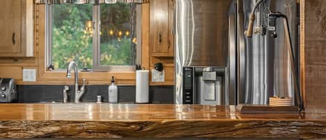 A wooden kitchen with a stainless steel refrigerator, a coffee machine, and a window with a view of greenery.