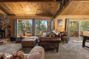 A cozy cabin interior with a stone fireplace, leather sofas, and a deck with outdoor furniture. - Heavenly Bay Retreat | Hot Tub, Lake View & Games! (Big Bear Lake)