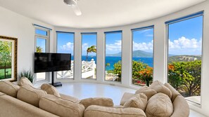 Un salon moderne doté d’un grand téléviseur à écran plat, de canapés beiges et offrant une vue panoramique sur l’océan grâce à de grandes fenêtres.