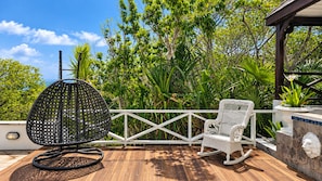 A wooden deck with a wicker chair and a rocking chair, surrounded by lush greenery and a white fence.