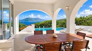 Ein runder Holztisch mit sechs Stühlen, einer verzierten Muschel und Blick auf Pool und Grünfläche.