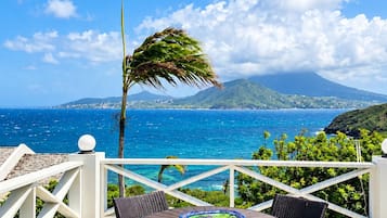 A terrace with wicker furniture and a view of the ocean and a mountain.