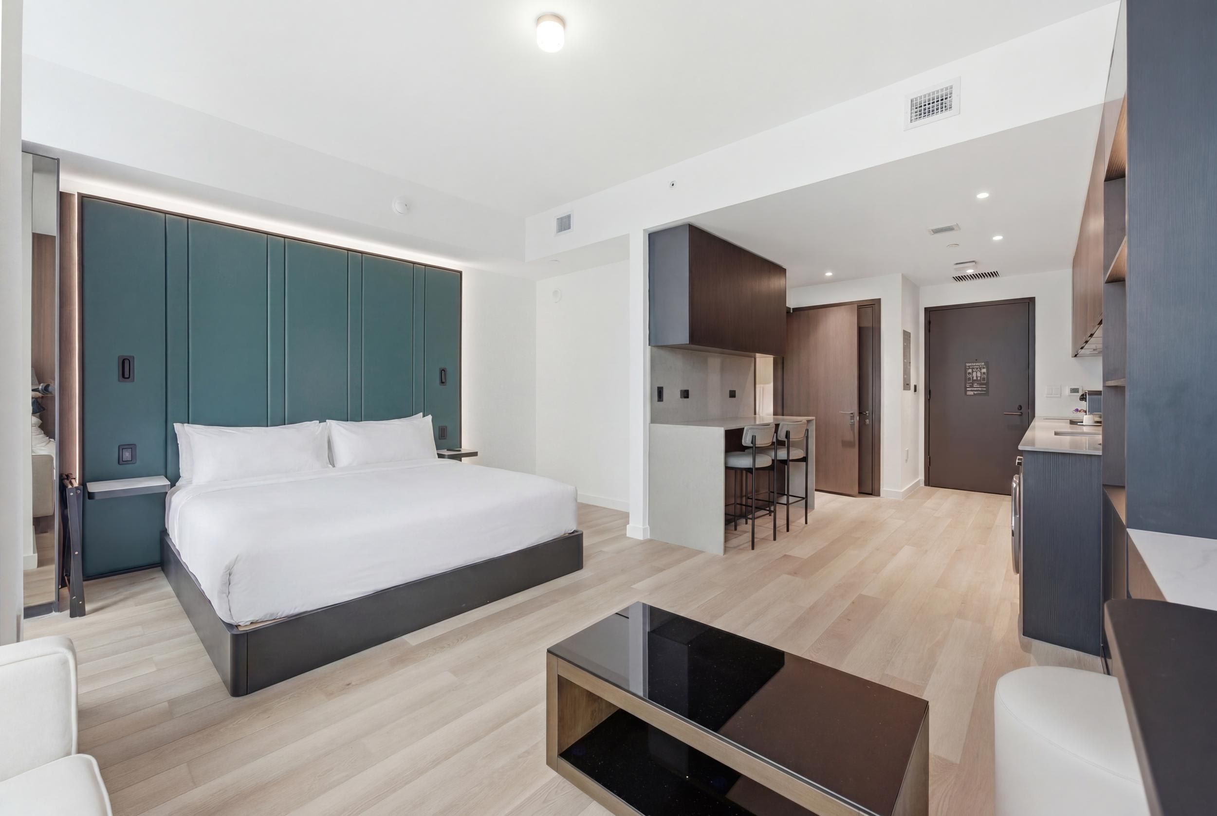A modern hotel room with a bed, a small table, a kitchenette, and a separate dining area.