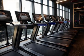 A row of treadmills with digital displays, positioned in a room with large windows offering a city view. - Luxury E11EVEN Suite with Amazing Pool & Views (Miami)