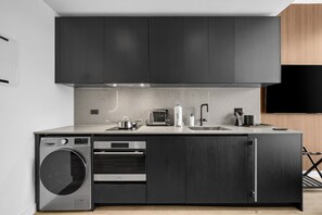 Une cuisine moderne avec des meubles de couleur sombre, un lave-linge encastré, une machine à café et un téléviseur à écran plat fixé au mur.