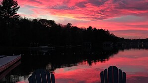 Ein Sonnenuntergangsblick von einer Anlegestelle mit zwei Adirondack-Stühlen, die auf einen ruhigen See ausgerichtet sind, umgeben von Bäumen und Gebäuden.