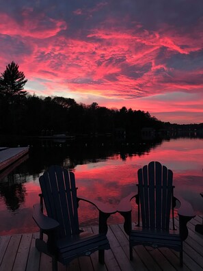 Vista del atardecer desde un muelle con dos sillas Adirondack mirando a un lago tranquilo, rodeado de árboles y edificios.