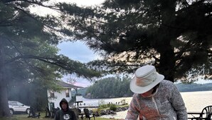 Eine Gruppe von Menschen versammelt sich um ein Lagerfeuer in einer Wiese, im Hintergrund sind ein See und Bäume zu sehen.