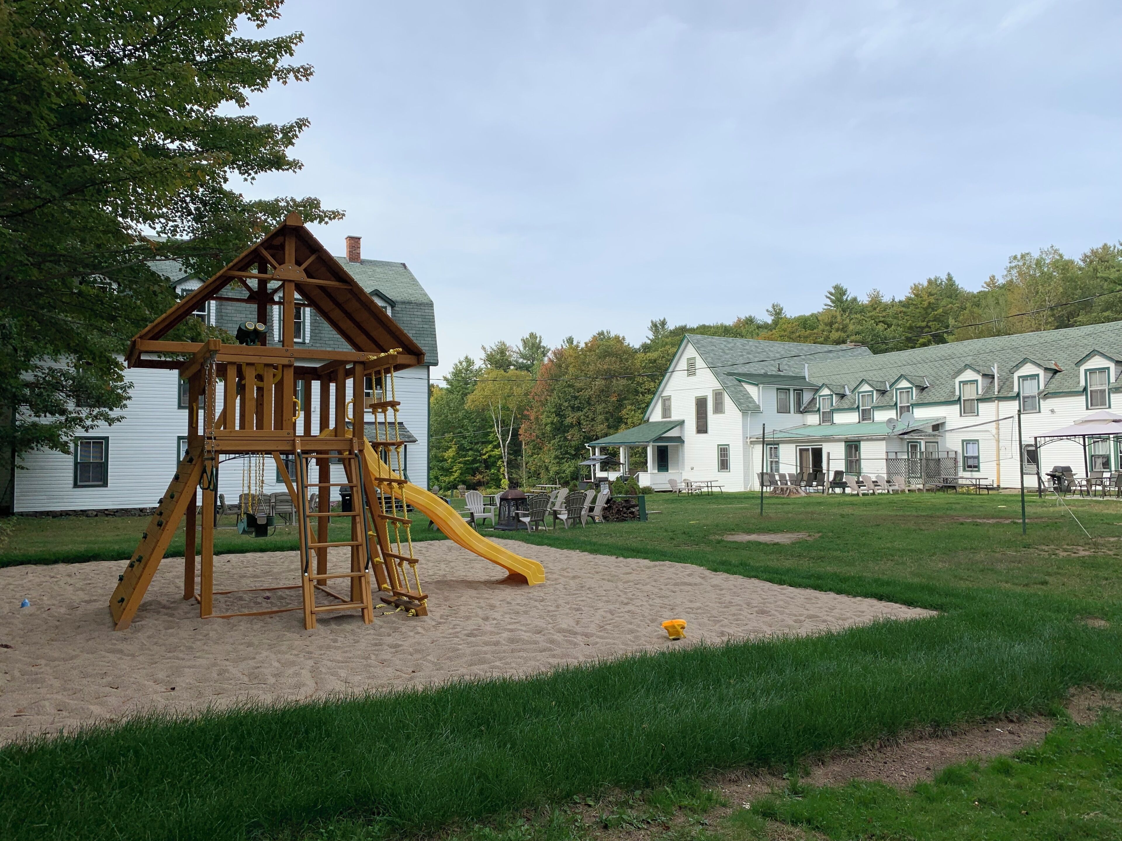 A playground with a wooden play structure and a slide, surrounded by a grassy area and a large white building with multiple windows and a green roof.