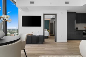 Een moderne woonkamer met een flatscreentelevisie, een eethoek met een tafel en stoelen, en een keuken met donkere kastjes en een koffieapparaat.