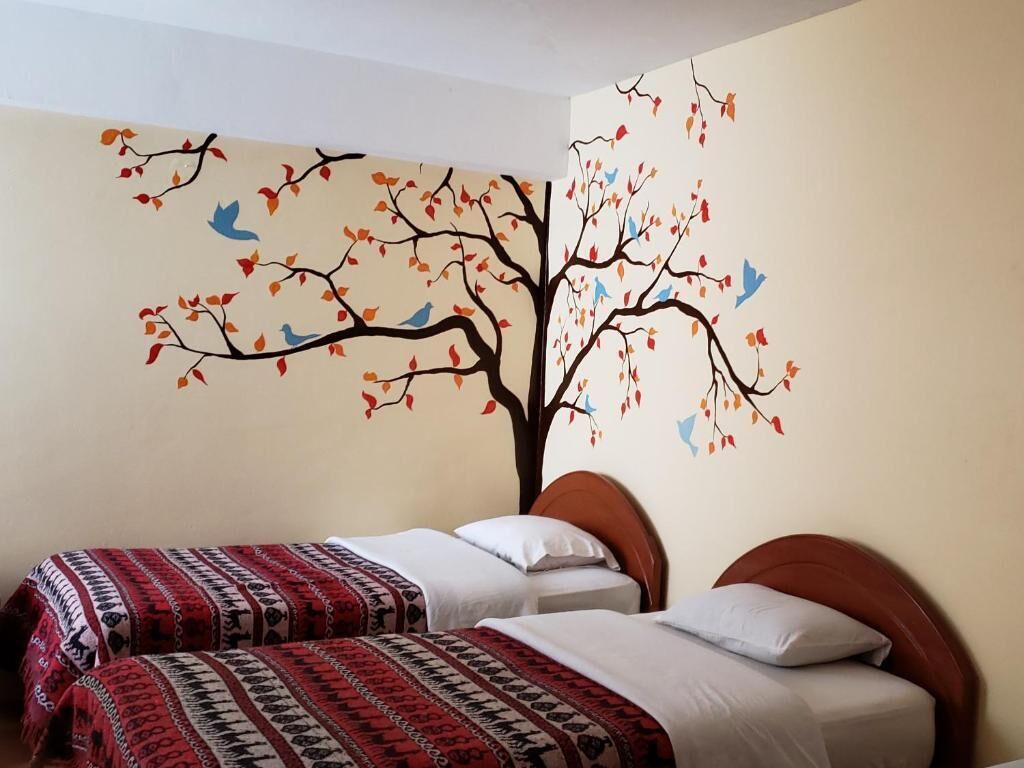 Un dormitorio con dos camas, un mural en la pared con un árbol, hojas y pájaros, y una pared lisa.