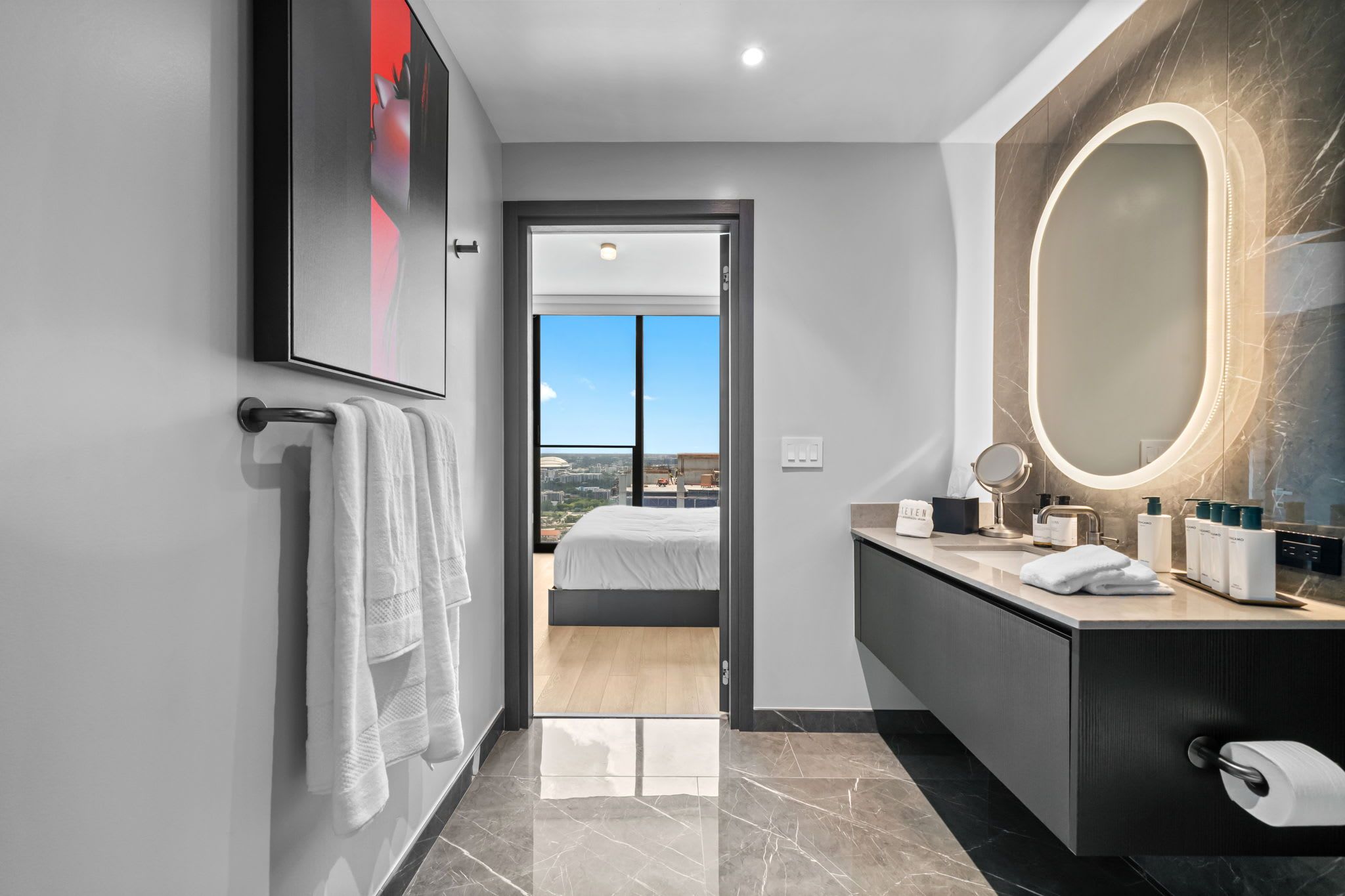 Une chambre d’hôtel moderne avec un grand lit, une salle de bain équipée d’un meuble-lavabo et d’un miroir, et une vue sur la ville depuis la porte.