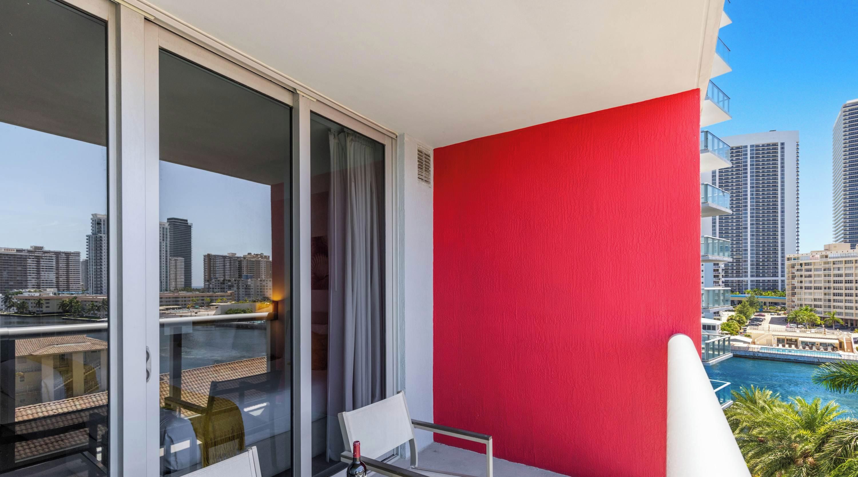 Un balcon avec un mur rouge, une porte vitrée et une vue sur le paysage urbain et l’eau.