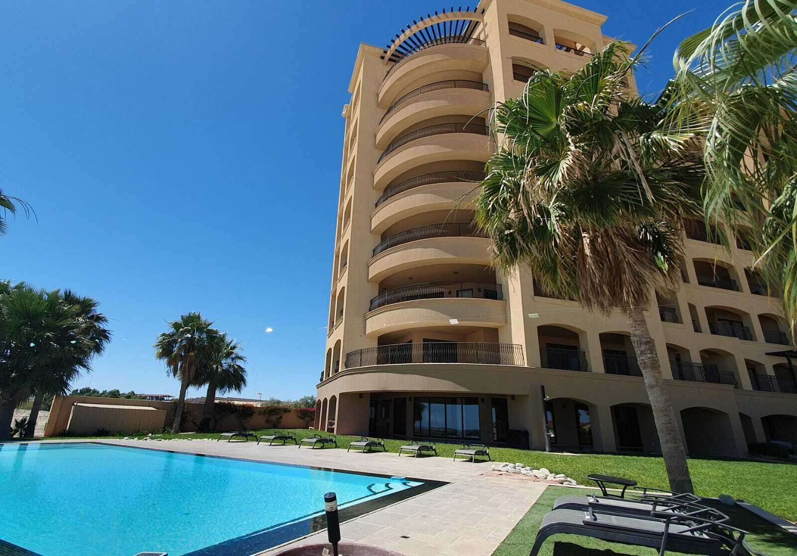 A multi-story hotel with a pool area, palm trees, and a clear blue sky.